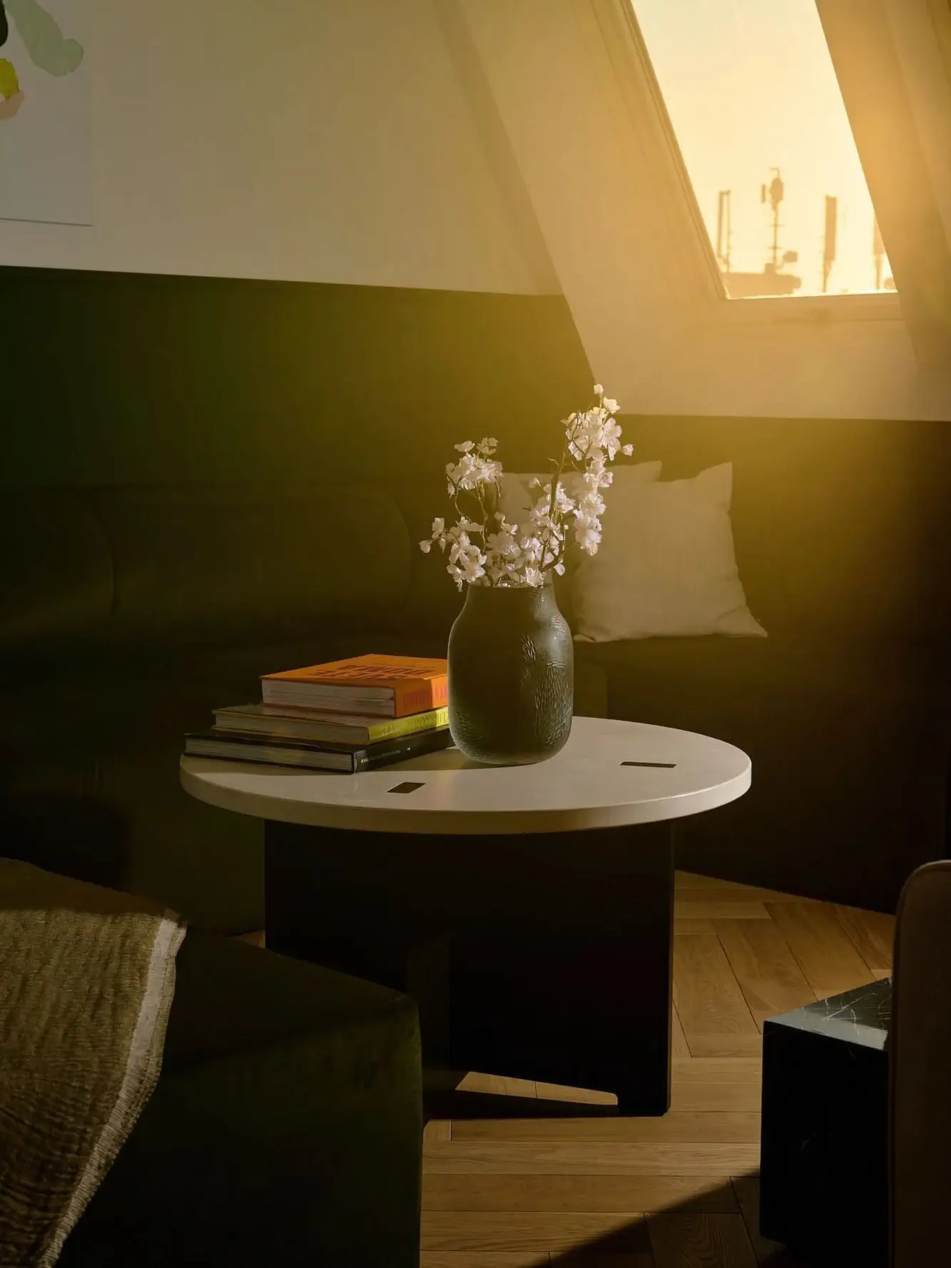 Ein runder weißer Couchtisch mit einer schwarzen Vase mit weißen Blumen und gestapelten Büchern steht im sonnendurchfluteten Wohnzimmer der PRÄSIDENTEN-SUITE, das durch ein dunkles Sofa und schräge Fenster akzentuiert wird.